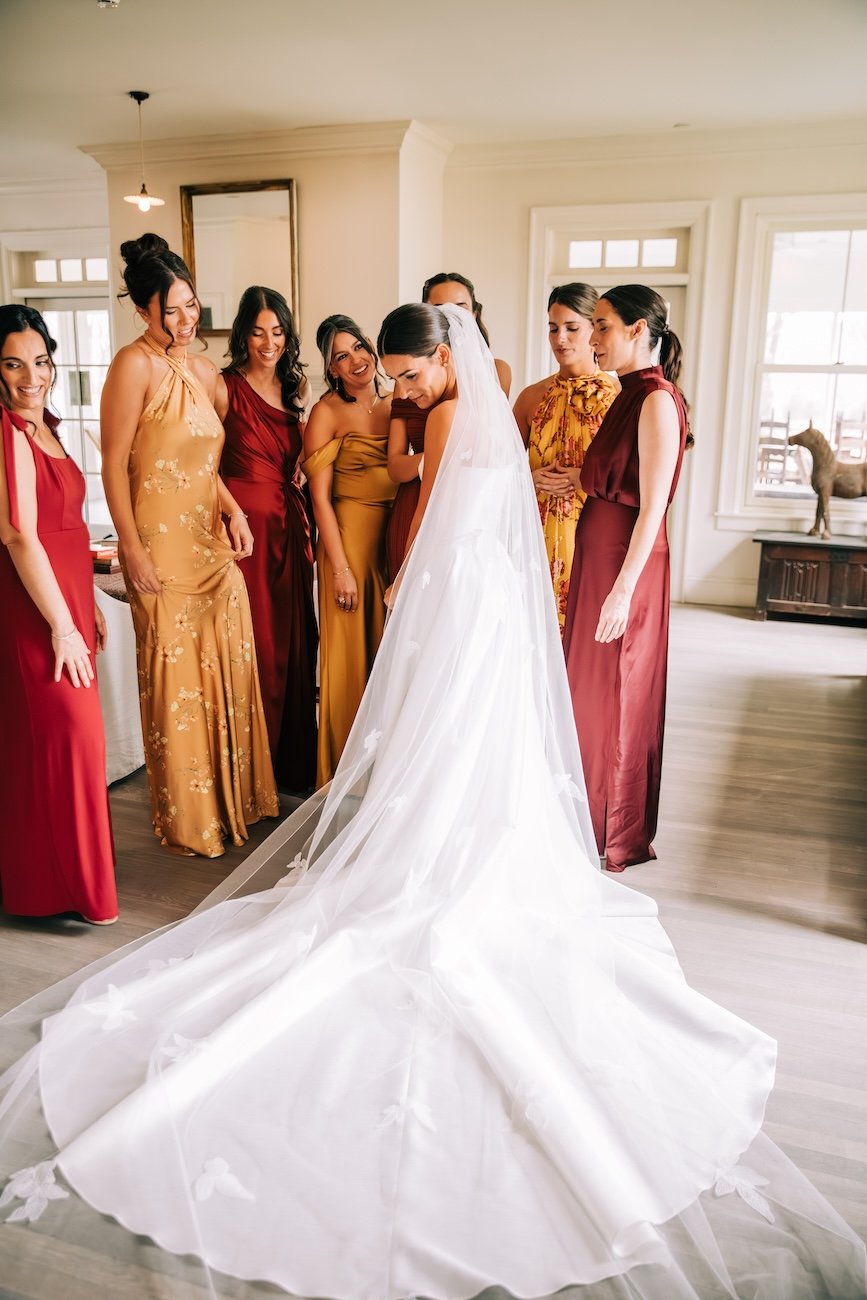 Bride in her wedding gown admiring her mom's repurposed cathedral veil with her bridesmaids wearing autumn colored gowns looking on.