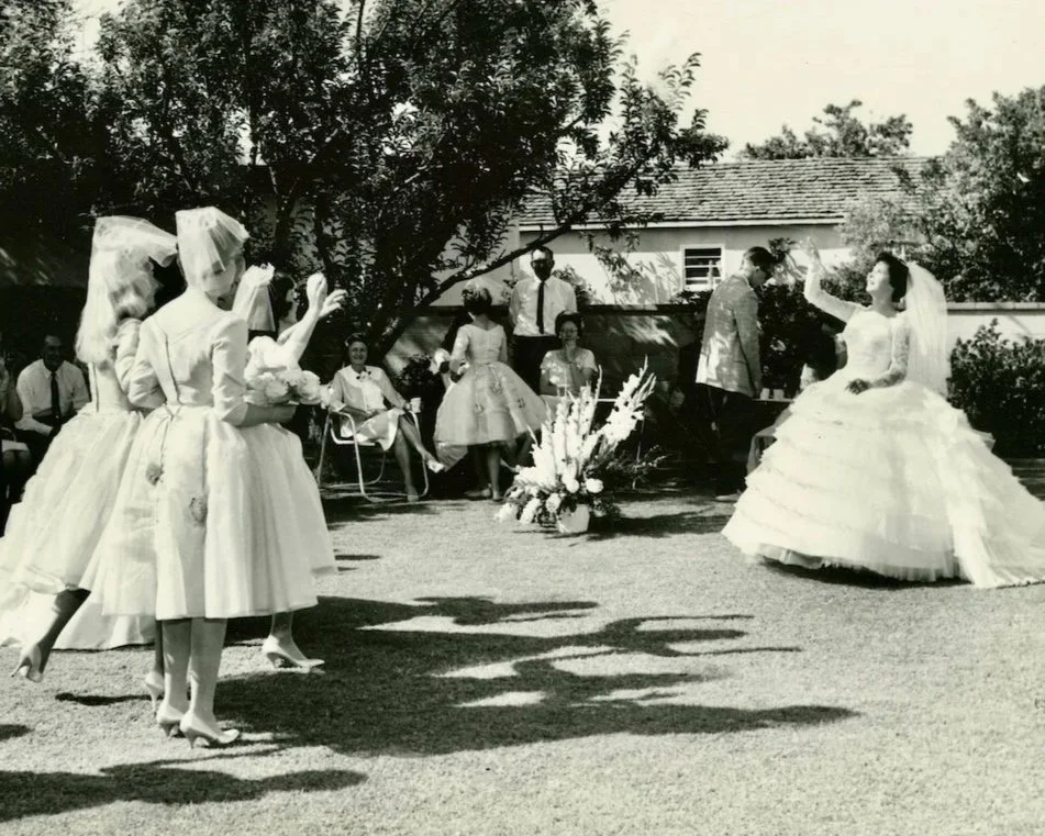 Bride tossing bouquet to bridemaids wearing flyaway veils at backyard wedding in 1961.