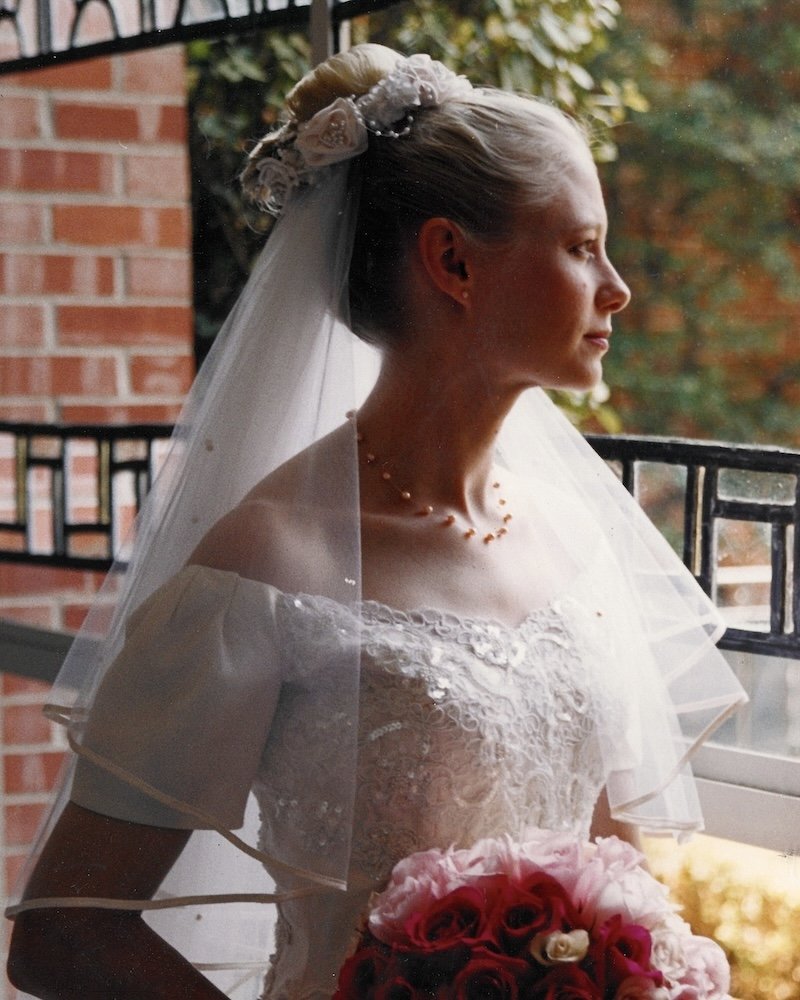 90s bride waering a blush colored satin floral bun wrap and satin trimmed wedding veil looking out a window.