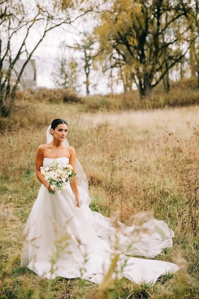 Gorgeous bride in an open field, pinching her cathedral veil slightly at her hip while holding her bouquet in the other hand.