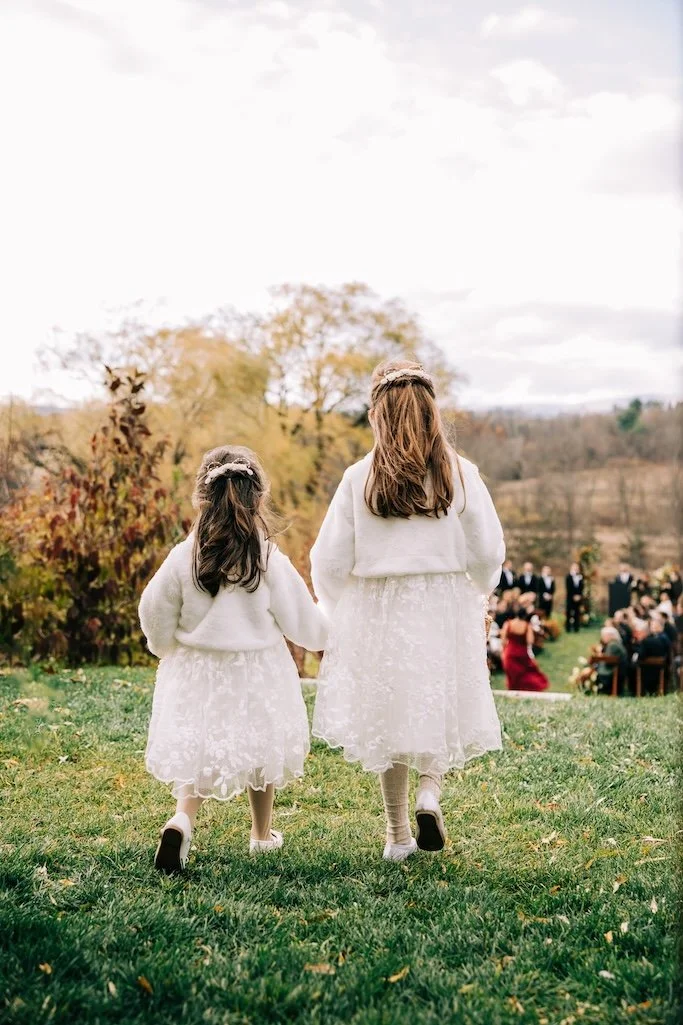 Two flower girls wearing white dresses and headpieces repurposed from a vintage bridal headpiece while walking down a grassy aisle towards wedding guests.