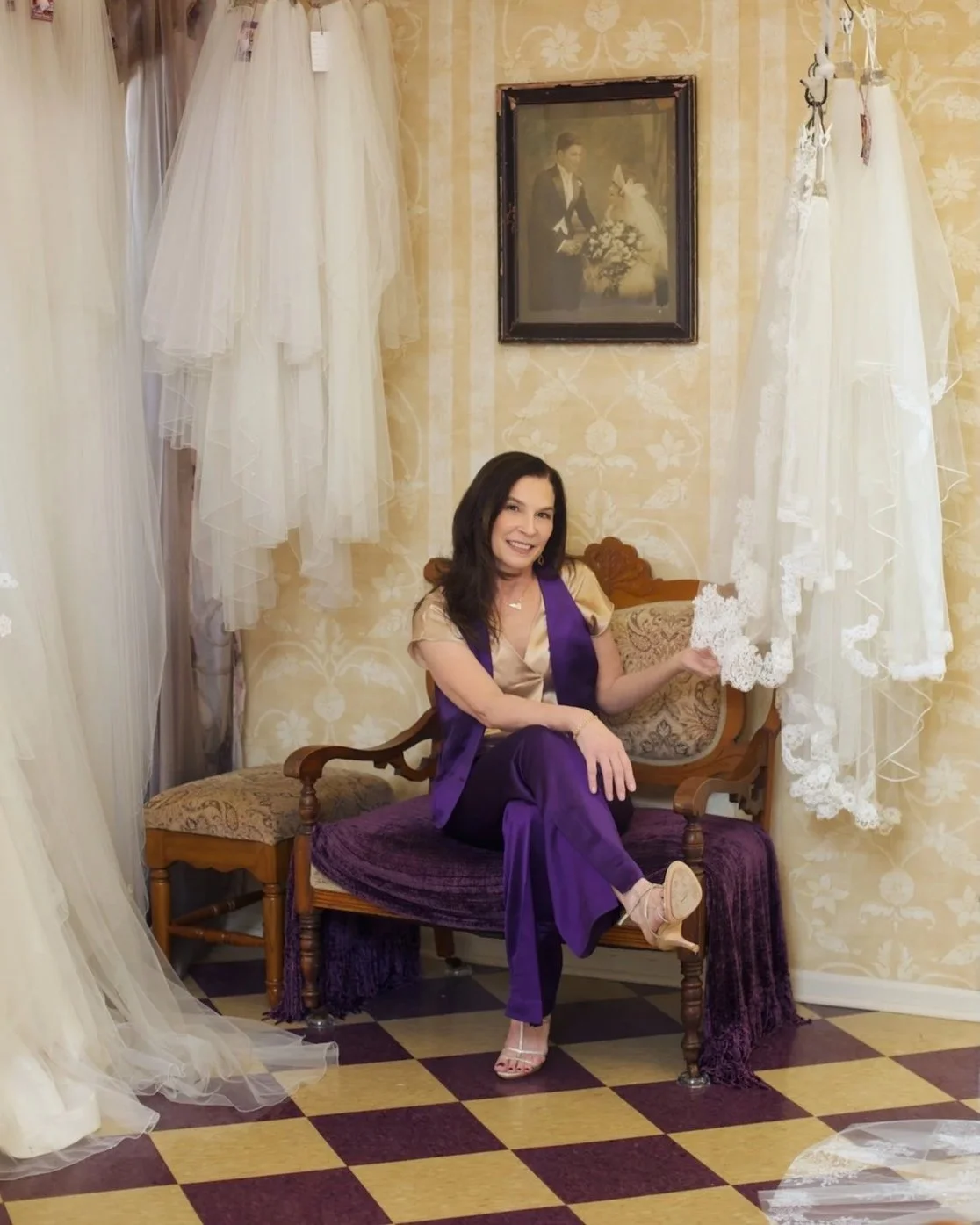 Designer, Marie Hunt, wearing her purple and gold signature colors, sitting in her shop touching a lace bridal veil.
