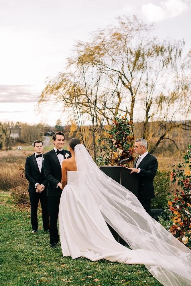 Bride's veil captured from behind, blowing in the wind during wedding vows on a beautiful fall day.