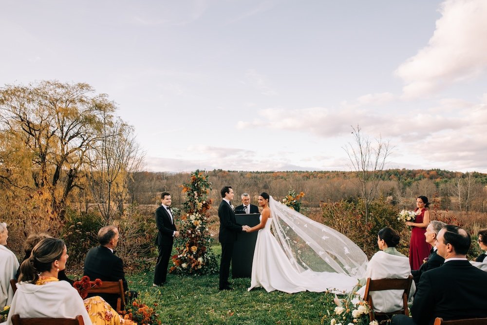 Beautiful outdoor autumn scene of bride and groom exchanging vows with her restyled long veil with leaf embroidery blowing in the wind.