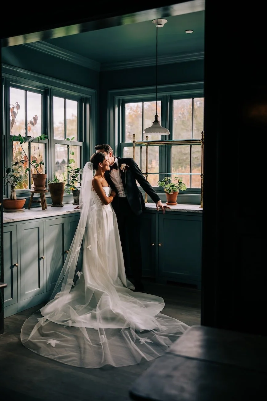 Bride and groom embracing in a darkened room of a farmhouse with natural lighting coming from the windows behind.