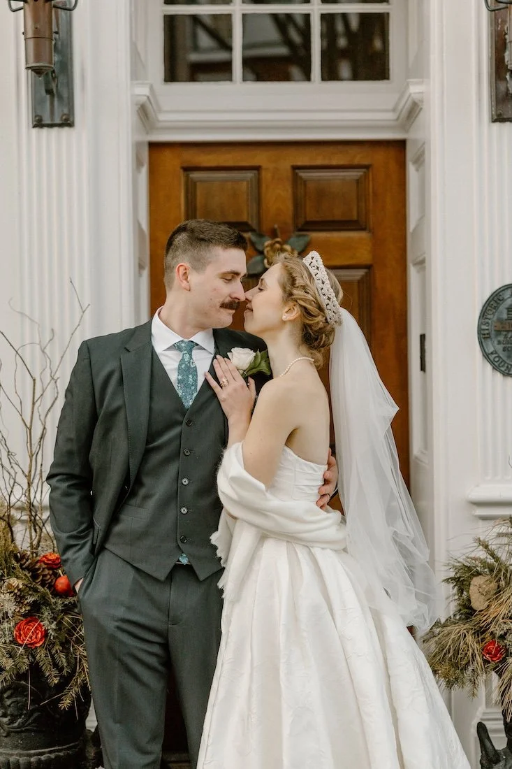 Groom and bride in front of white building with a wooden door nuzzling noses.