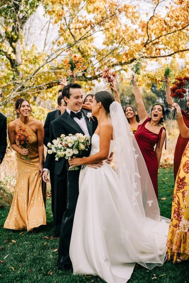 Bride and groom facing each other smiling under a tree with autumn leaves and their bridal party cheering them on.