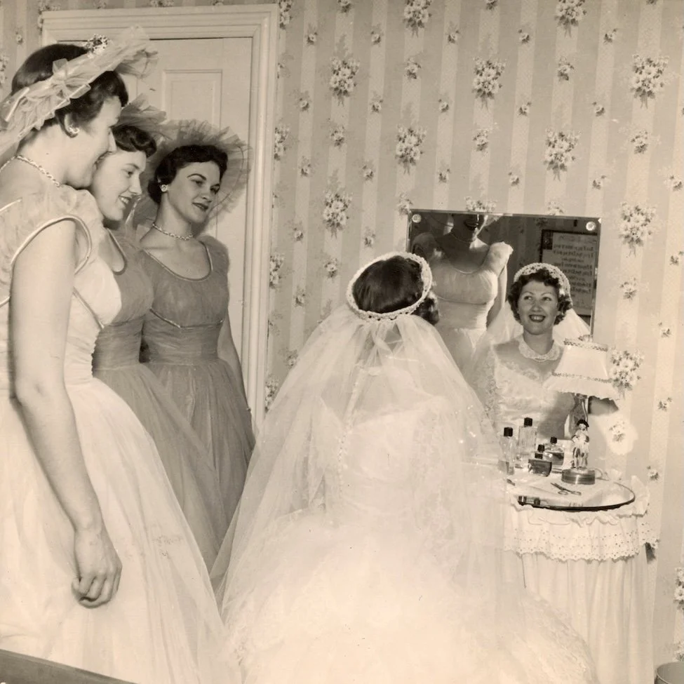 Vintage wedding photo of 50s bride looking in the mirror with her bridesmaids wearing bridal millinery, looking on.