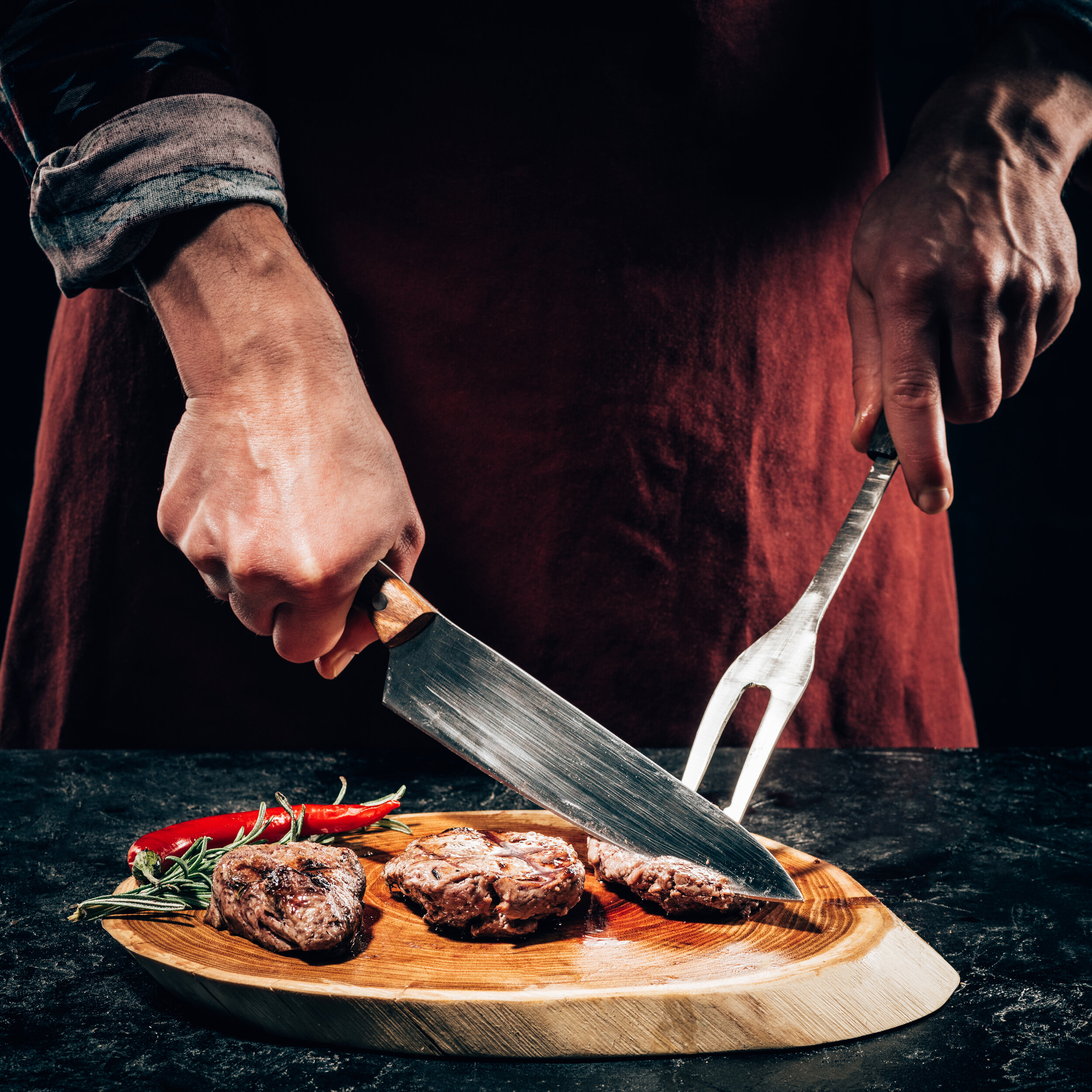 close-up-partial-view-chef-in-apron-with-meat-fork-and-knife-slicing-gourmet-grilled-steaks-with-rosemary-and-chili-pepper-on-wooden-board-902944120_4766x4766.jpeg