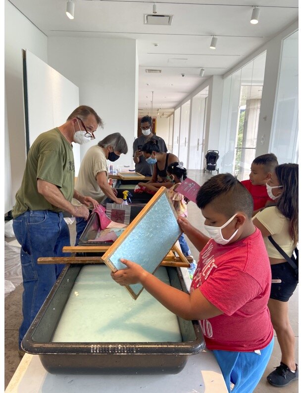 Youth in red T-shirt holding paper mould with blue pulp in foreground, with instructors and other students in background