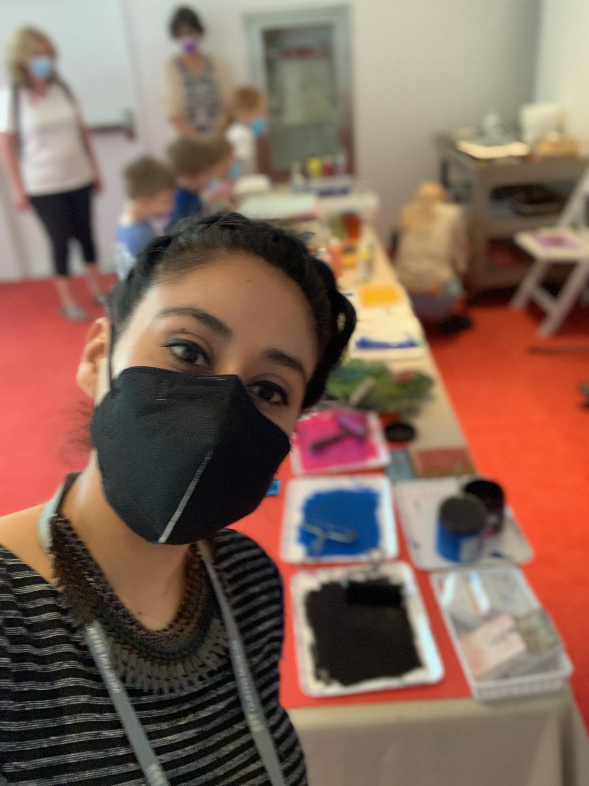 Selfie of Niceli in front of table with paint, wearhing black mask, striped shirt and ornate necklace