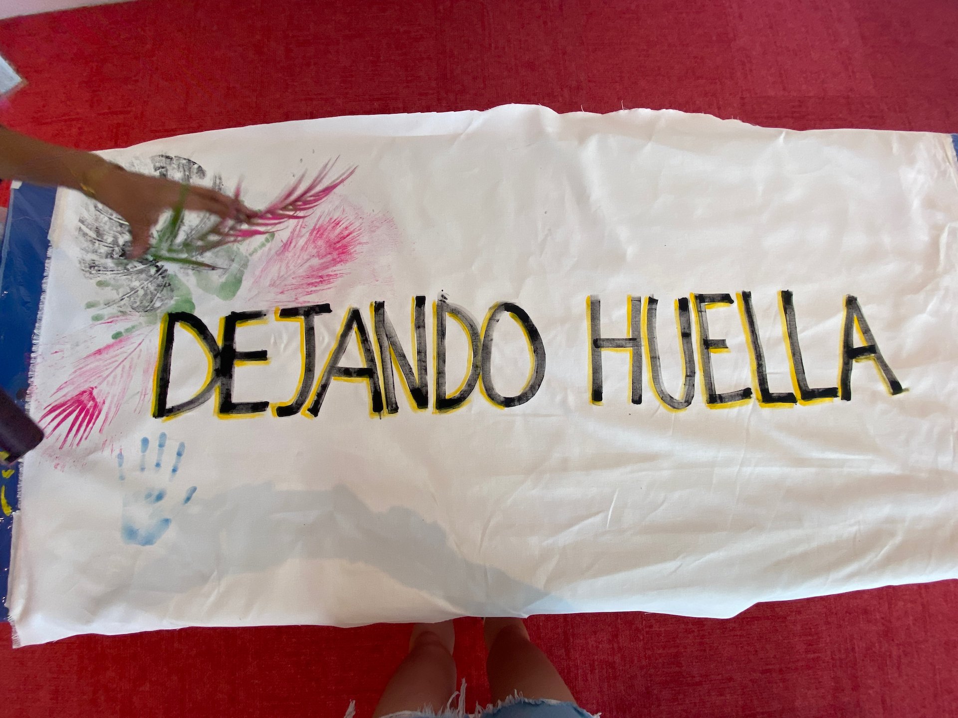 Long, white fabric with plant and hand prints, with hand-written black and yellow text that reads "Dejando Huella"