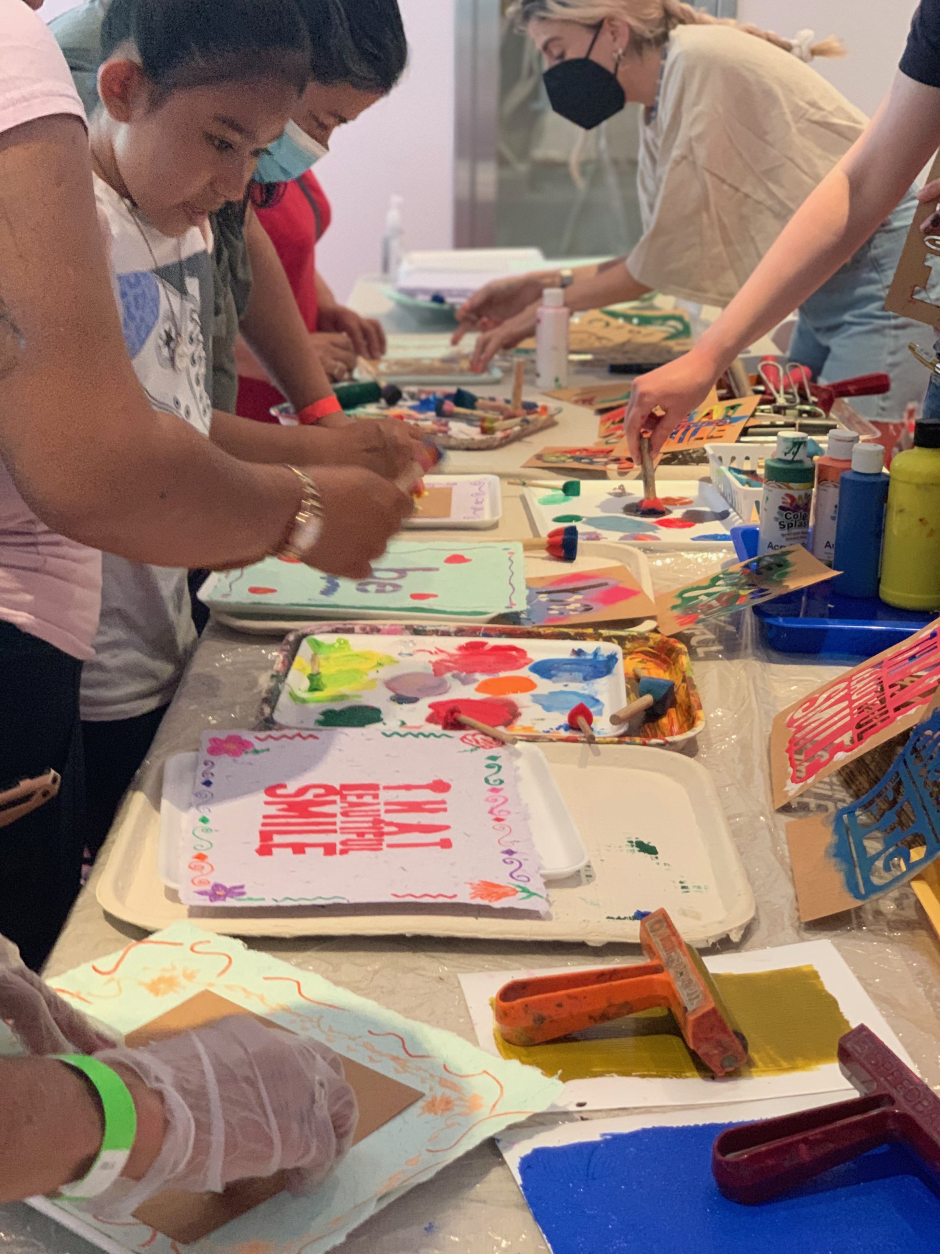 Camila working in background with familes at table, stenciling words on mulitcolored handmade paper. Closest stencil reads "that beautiful smile" with floral border