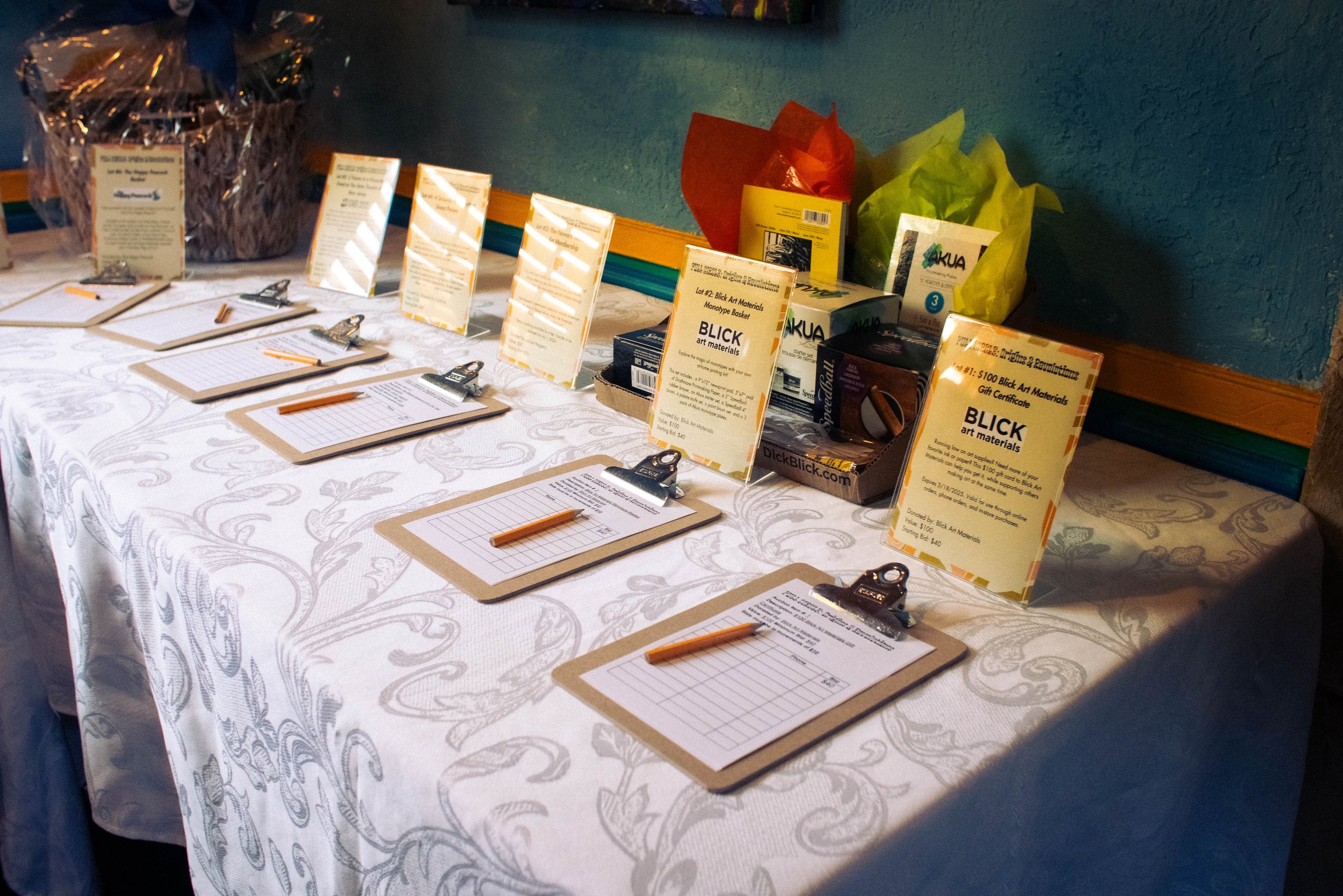  Five clipboards with auction sign-ups are displayed in a row, placed atop a cloth table with each one accompanied by a displayed pamphlet of each auction prize. 