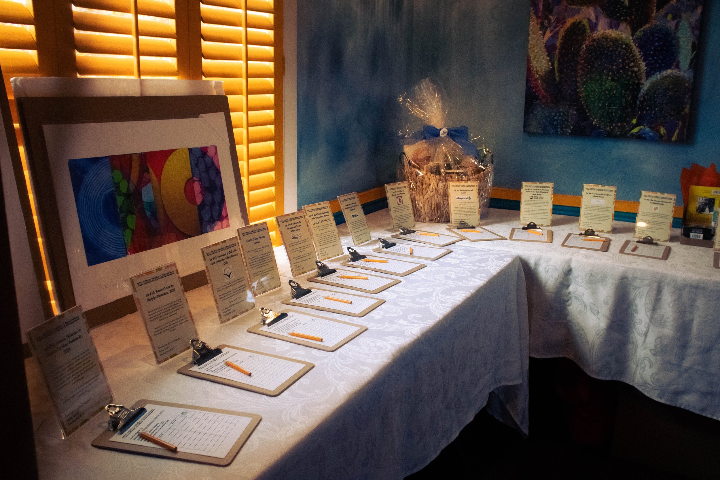  Several clipboards with auction sign-ups are displayed in a row, placed atop a cloth table with each one accompanied by a displayed pamphlet of each auction prize. A large colorful print is placed on the window in an presentable display. 