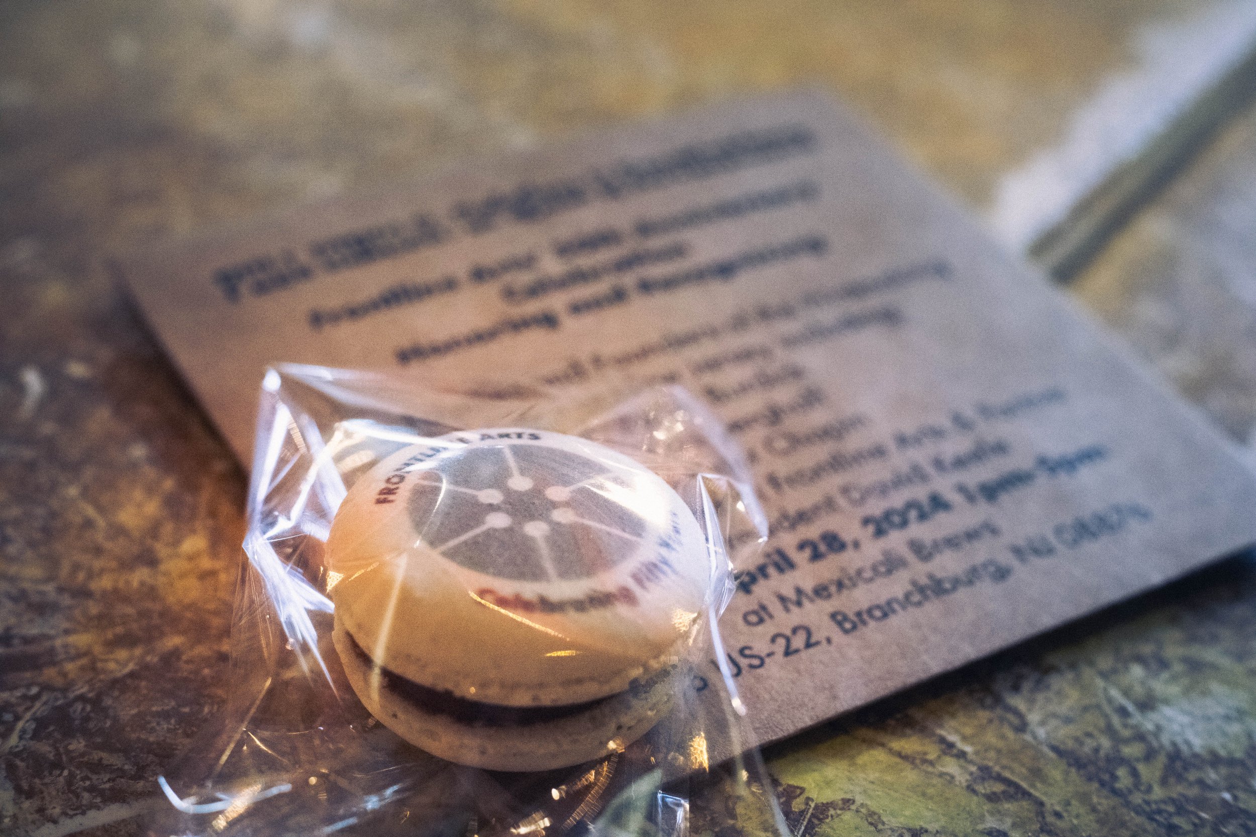  A sealed macaron with the Frontline Arts logo printed on top of it lays on a table with an itinerary laying underneath like a place mat. The itinerary describes the event and honorees. 