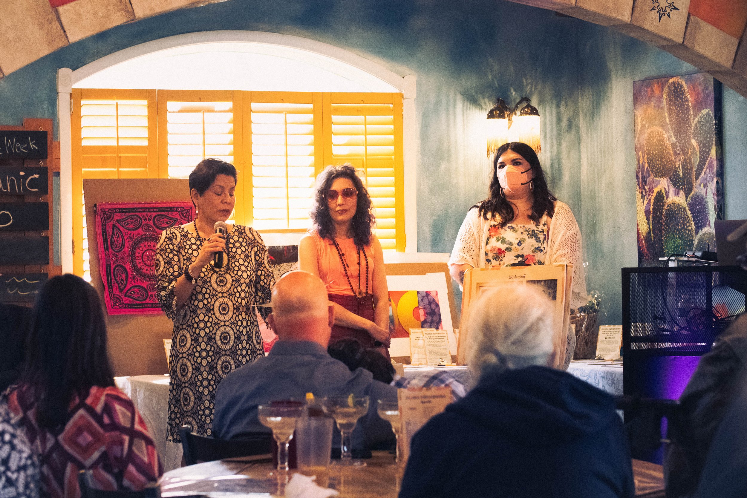  In front of the auction items displayed, Gianna Seaman, Rachel Heberling, and Lindsey Knipe are addressing the attendees while standing side-by-side as the window light illuminates the scene. 