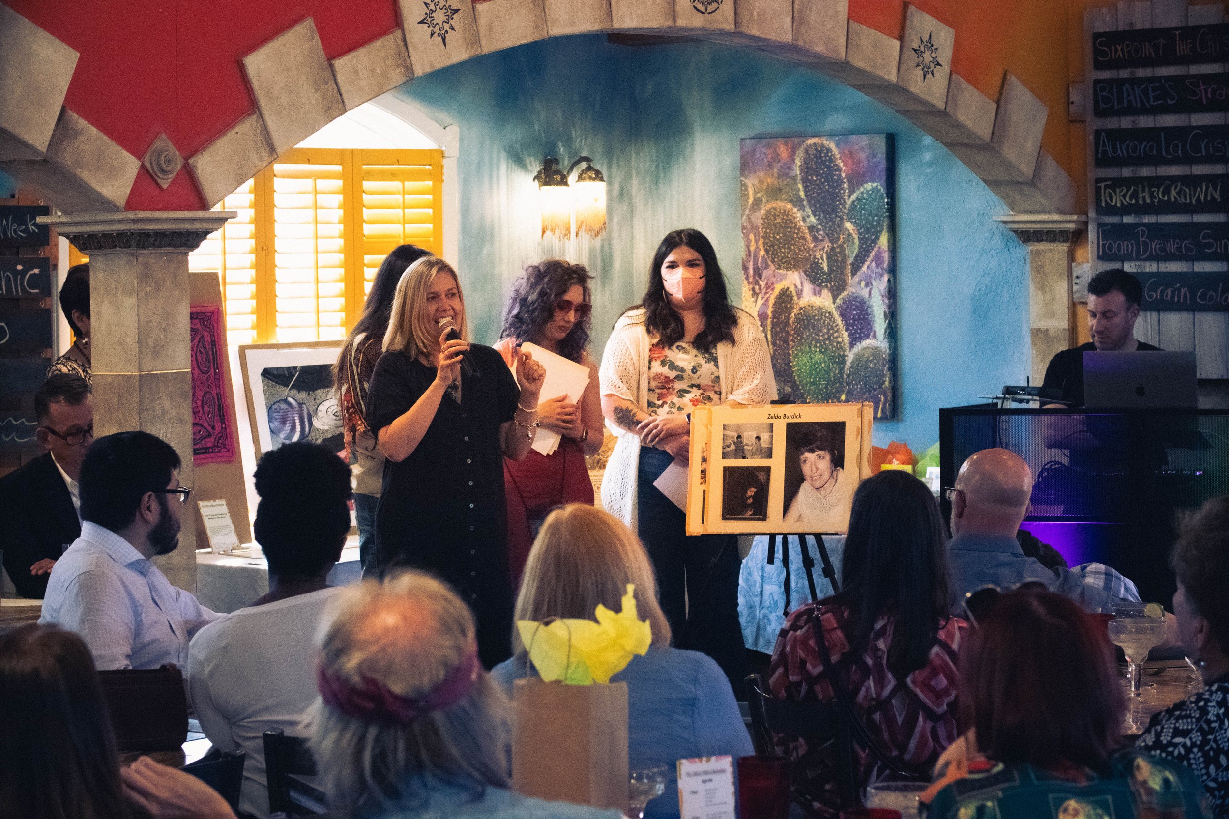  In front of the auction items displayed, Gianna Seaman, Rachel Heberling, and Lindsey Knipe are standing side-by-side as family member of Zelda Burdick (Shanna Hospelhorn) is addressing the seated attendees through a microphone. 