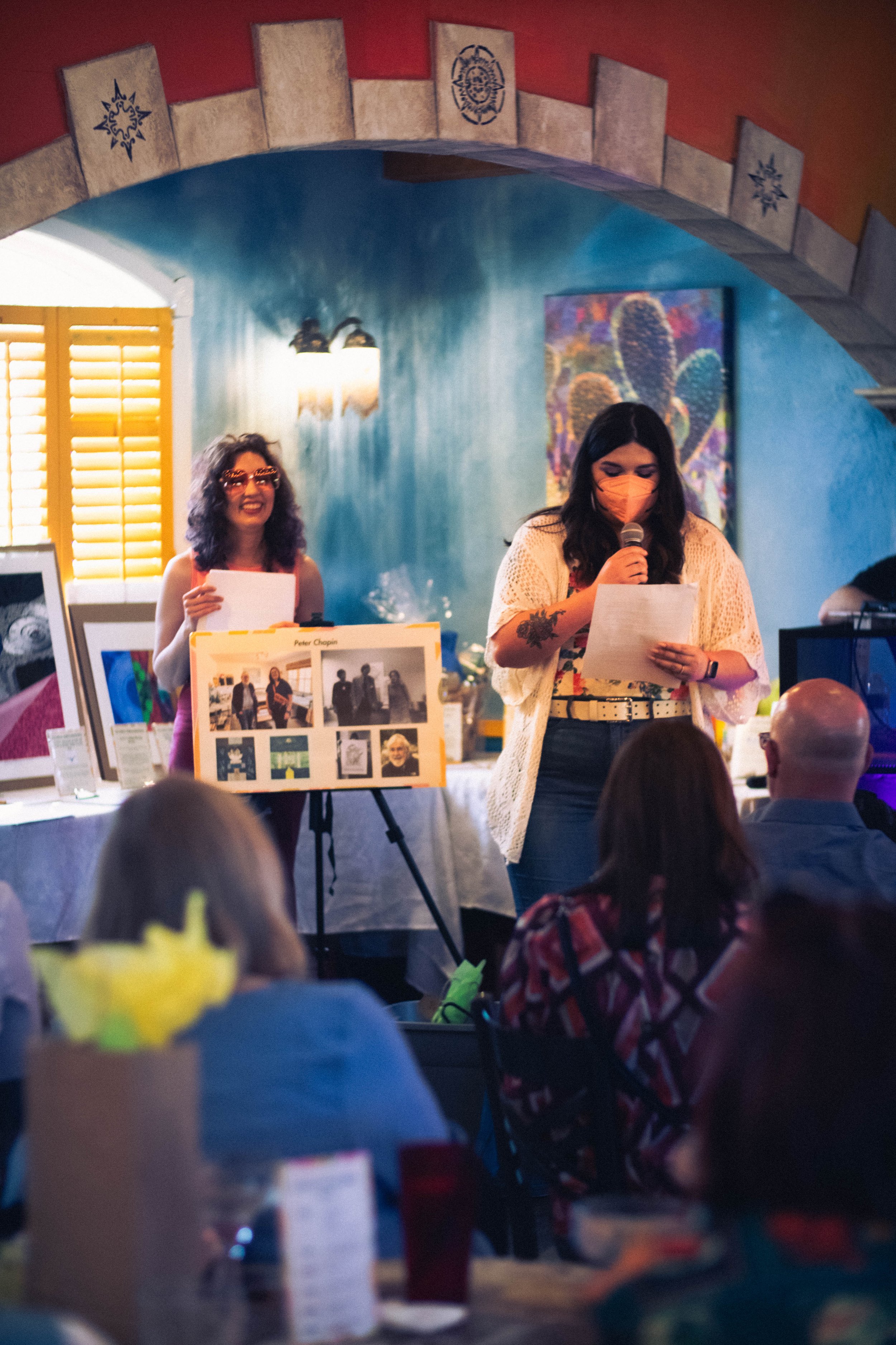  In front of the auction items displayed, Rachel Heberling and Lindsey Knipe are presenting highlights of honorees, with Rachel holding a display of pictures arranged in a collage and Lindsey reading from a paper as she talks. 