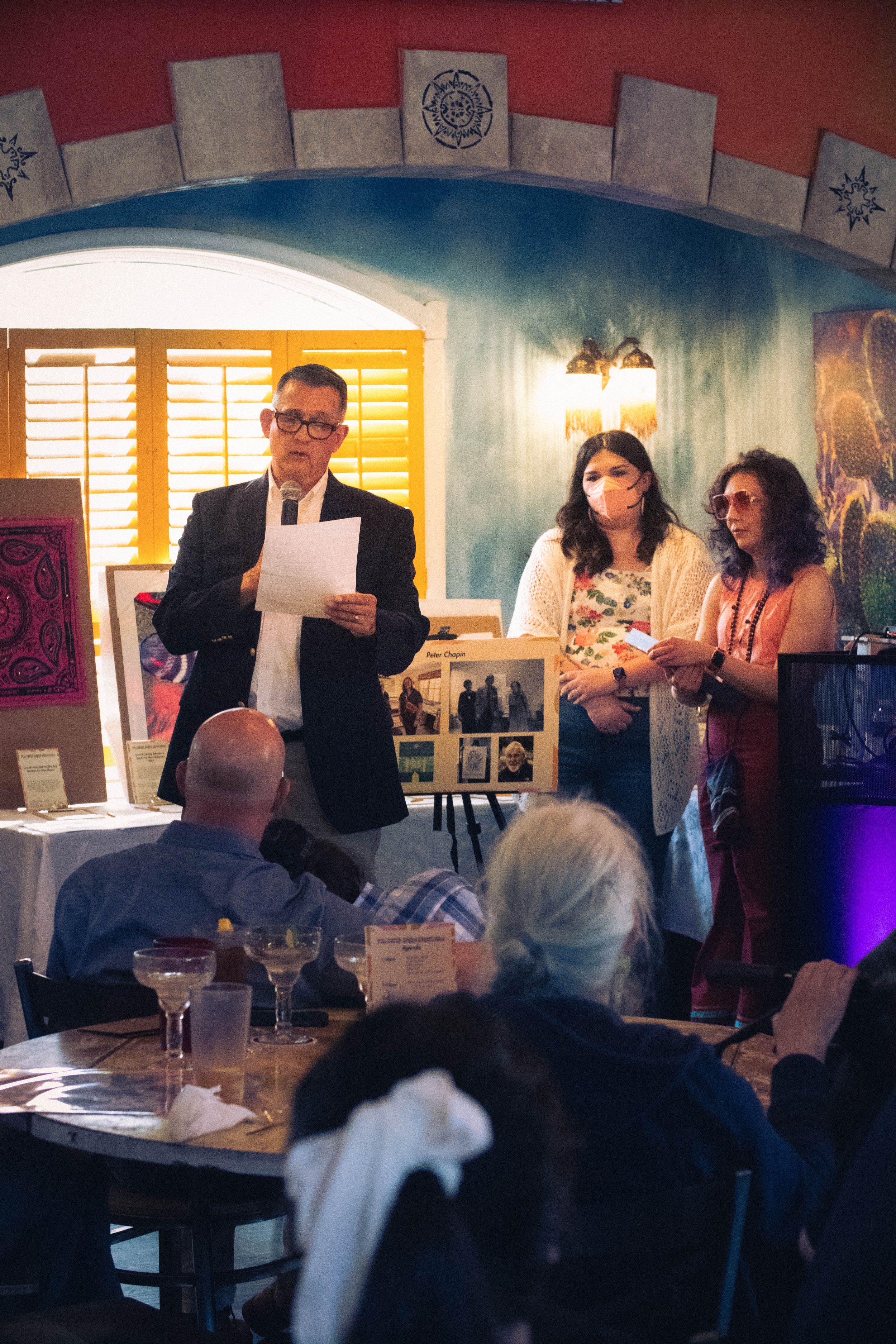  In front of the auction items displayed, Ron Erickson, Rachel Heberling, and Lindsey Knipe are addressing the attendees while standing side-by-side as the window light illuminates the scene. 