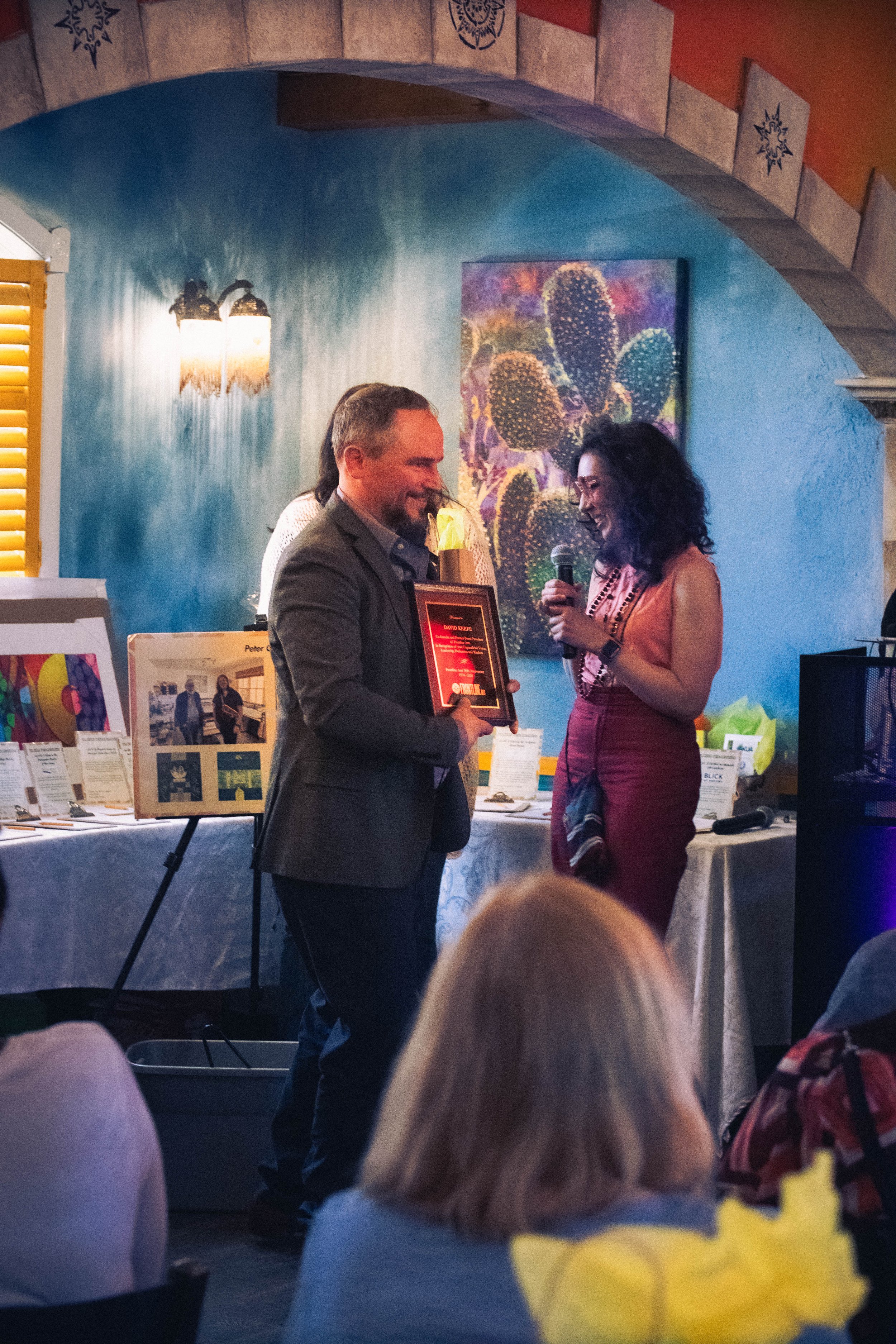  Dave Keefe holds his awarded plaque of wood and golden borders as he smiles to the attendees, with Rachel Heberling standing close by in front of the table with displayed auction items. 