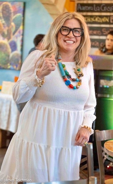  At attendee with colorful necklaces, a white dress and blonde hair smiles for the camera as she poses for a photograph. 