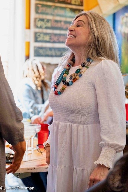  At attendee with colorful necklaces, a white dress and blonde hair smiles as she's in conversation with another off-screen. 