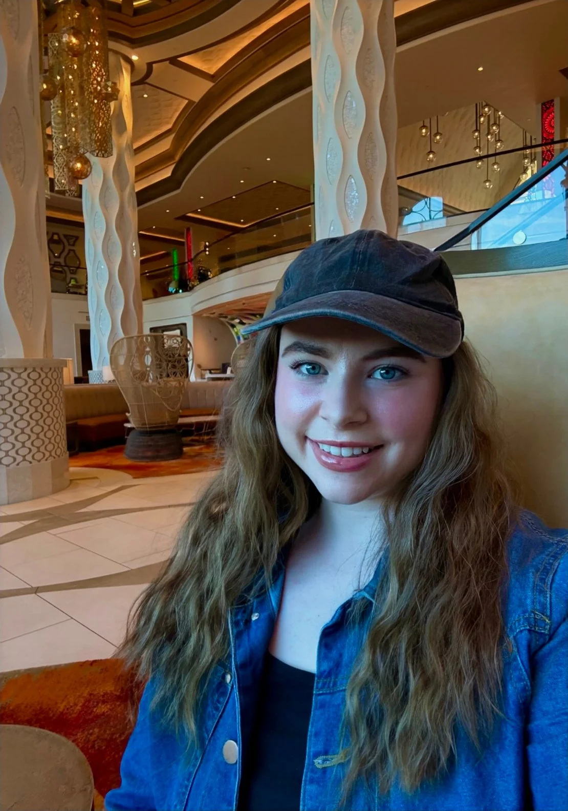 A young woman with long wavy hair wearing a black cap, black top, and denim jacket, smiling while sitting in a modern hotel lobby with warm lighting and decorative elements.