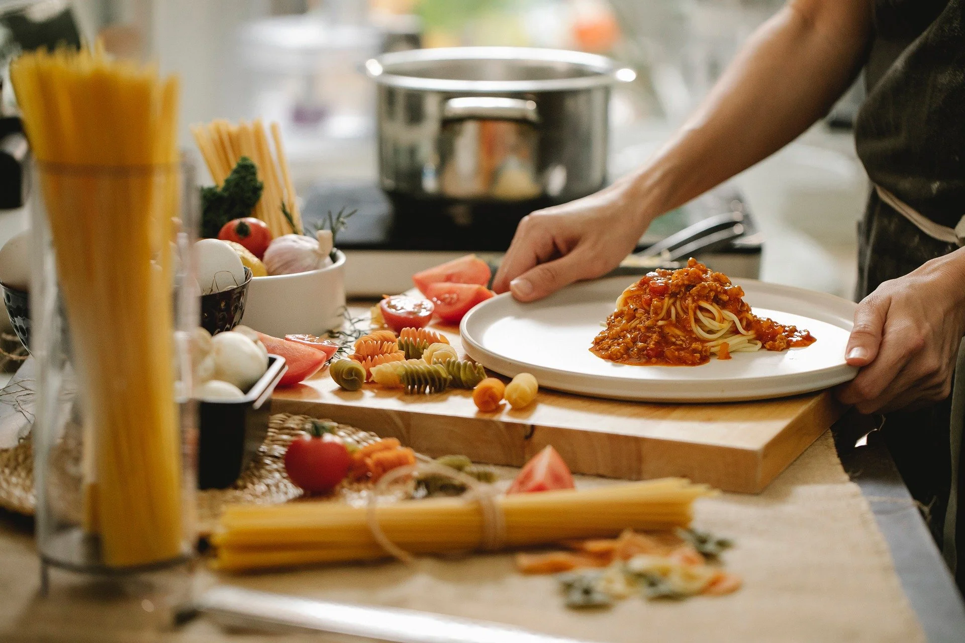 Person plating spaghetti with meat sauce in a kitchen surrounded by fresh vegetables and pasta.