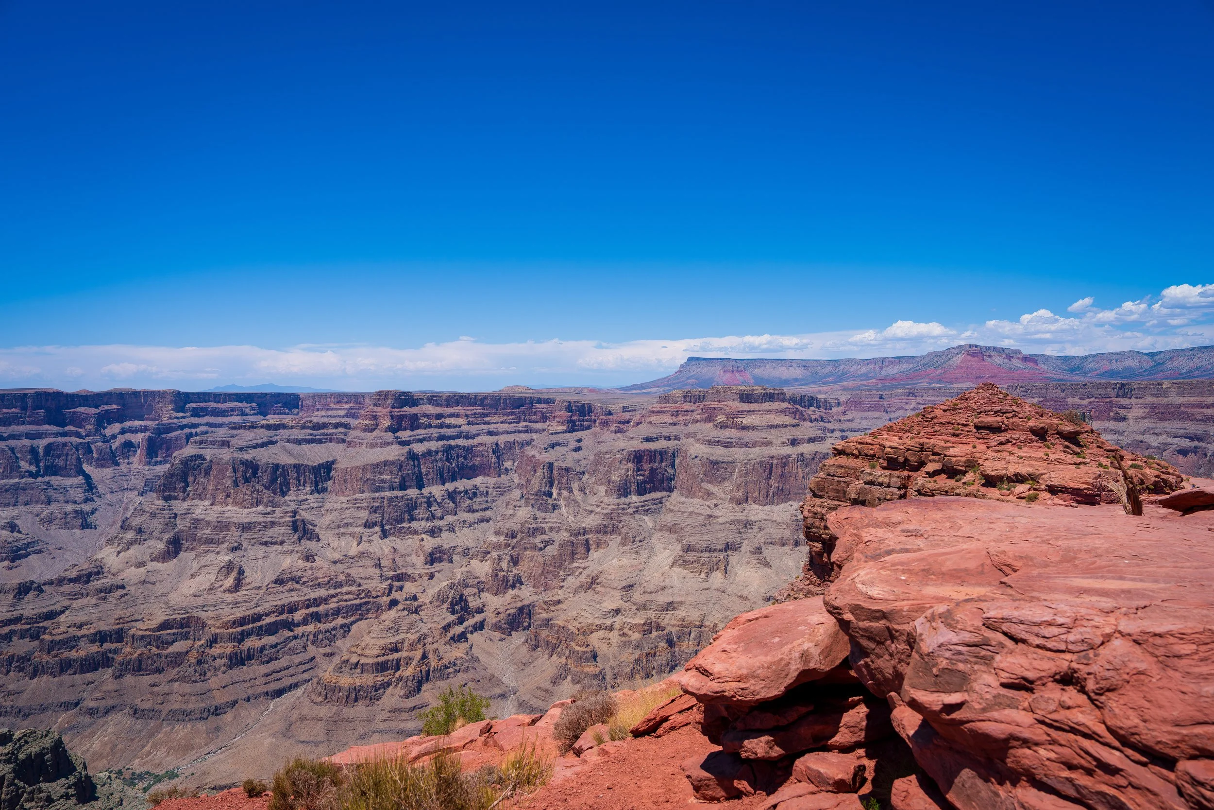 A panoramic view of the Grand Canyon under a bright blue sky with a few scattered clouds, showcasing layered rock formations and rugged terrain.