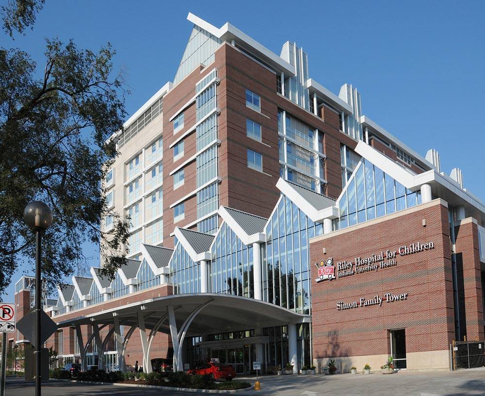A modern multi-story hospital building with red brick and glass facade. Signage indicates it is the Riley Hospital for Children, part of Indiana University Health, named the Simon Family Tower. The entrance has a curved canopy with supporting arches, and there are cars parked in front.