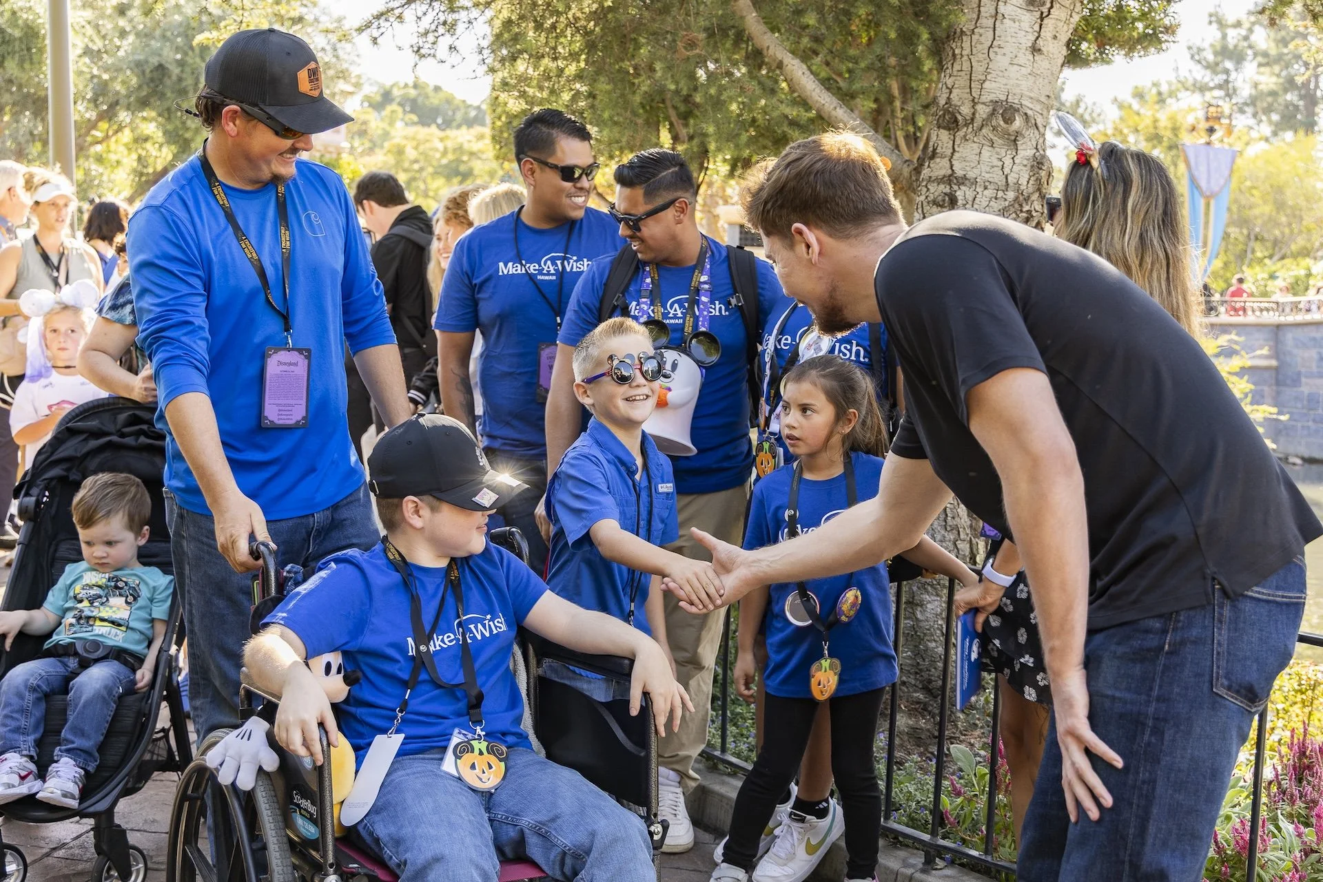Group of children and adults at an outdoor event, some wearing Disney-themed costumes and lanyards, with children in wheelchairs, shaking hands and smiling.