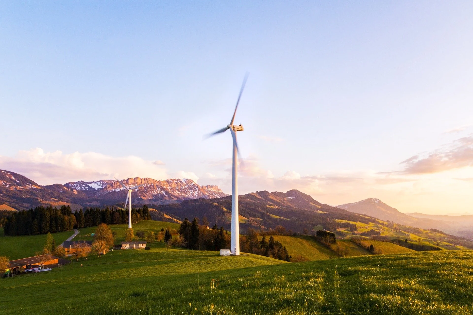 Scenic landscape with wind turbines on green rolling hills, mountains with snow patches in the background, and a partly cloudy sky at sunset.