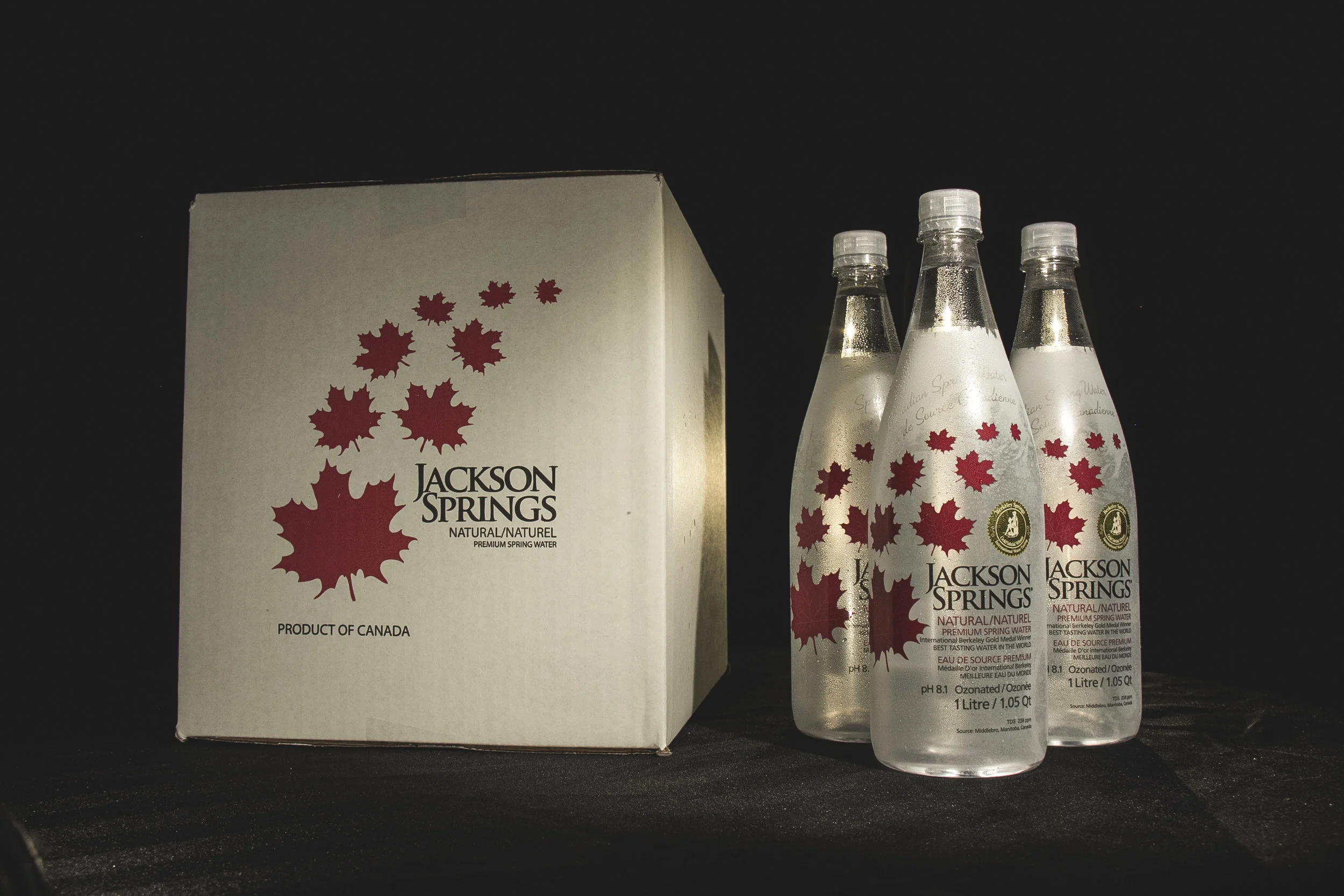 A cardboard box of Jackson Springs natural spring water and three glass bottles of Jackson Springs spring water with red maple leaf designs, set against a black background.