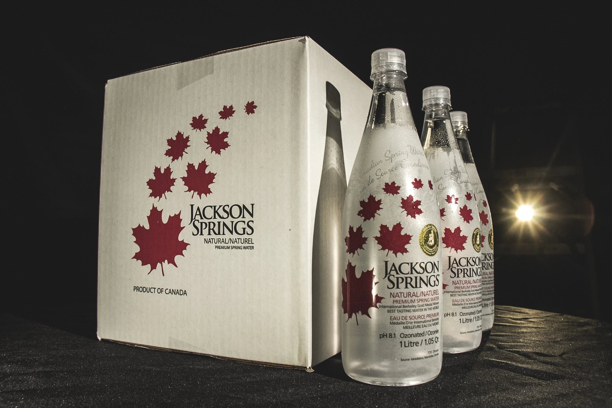 Three bottles of Jackson Springs sparkling water next to a white box with red maple leaf designs, on a dark surface at night