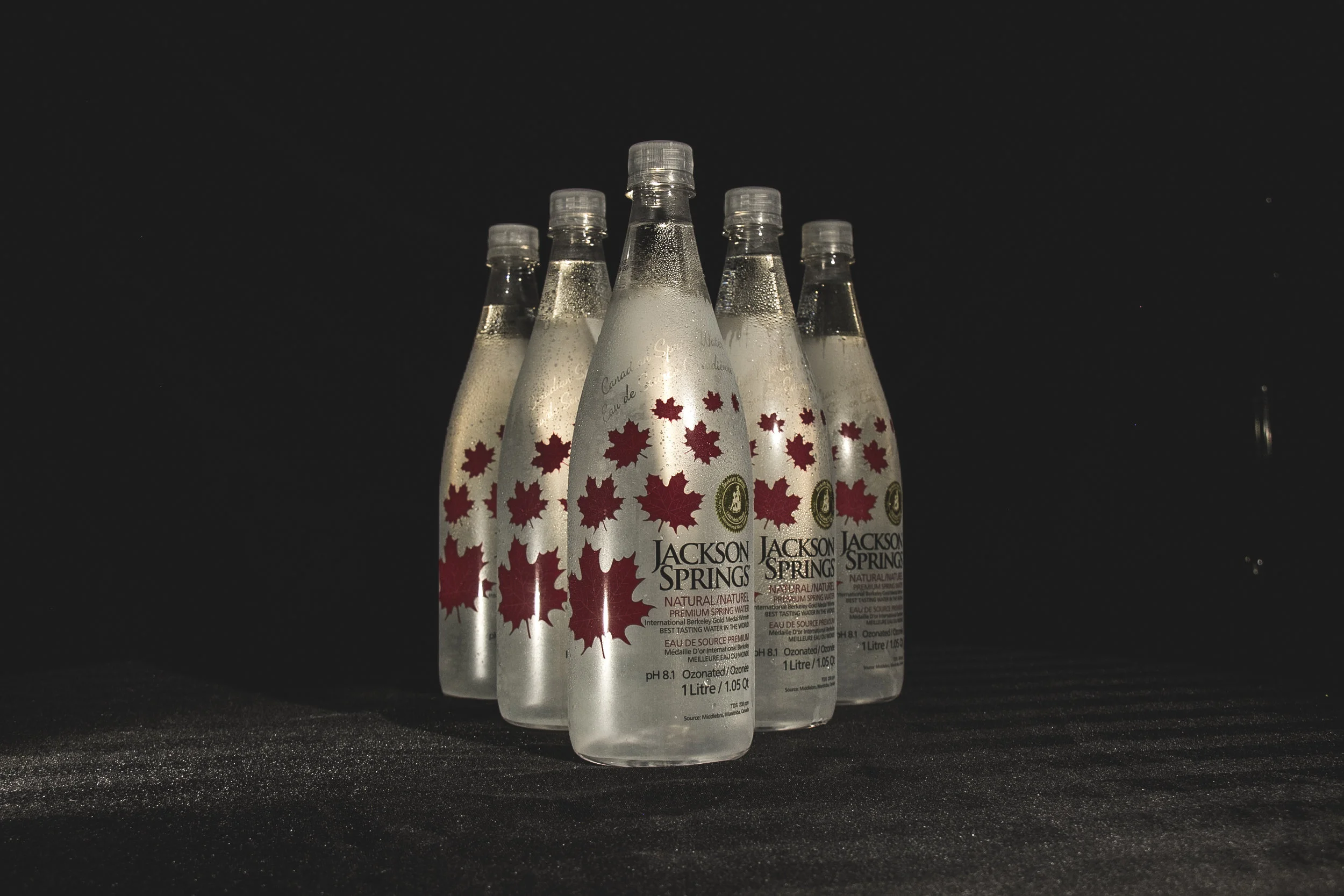 Six bottles of Jackson Springs bottled spring water with maple leaf graphics on a dark surface and background.