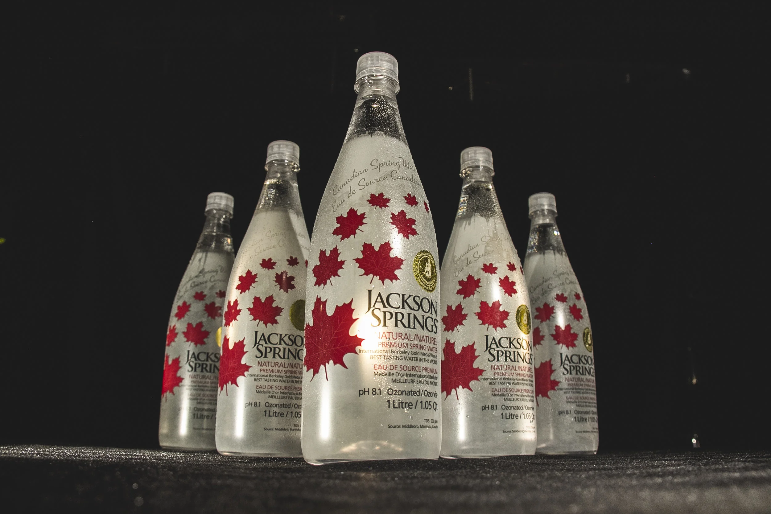 Five bottles of Jackson Springs natural spring water with red maple leaf designs on the labels, displayed on a dark surface against a dark background.