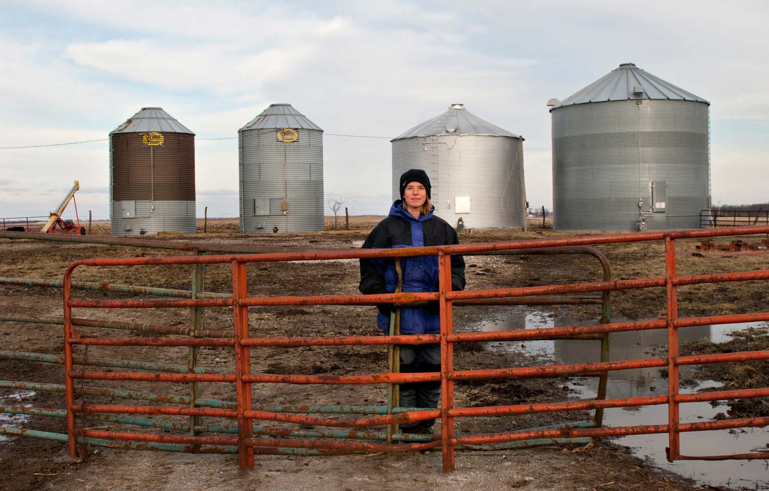  Kristy Bollin, farmer and wife standing at her family’s farm.  Orange metal fence and grain bins n the background. Countryside of Bowen, IL. March, 2009. 