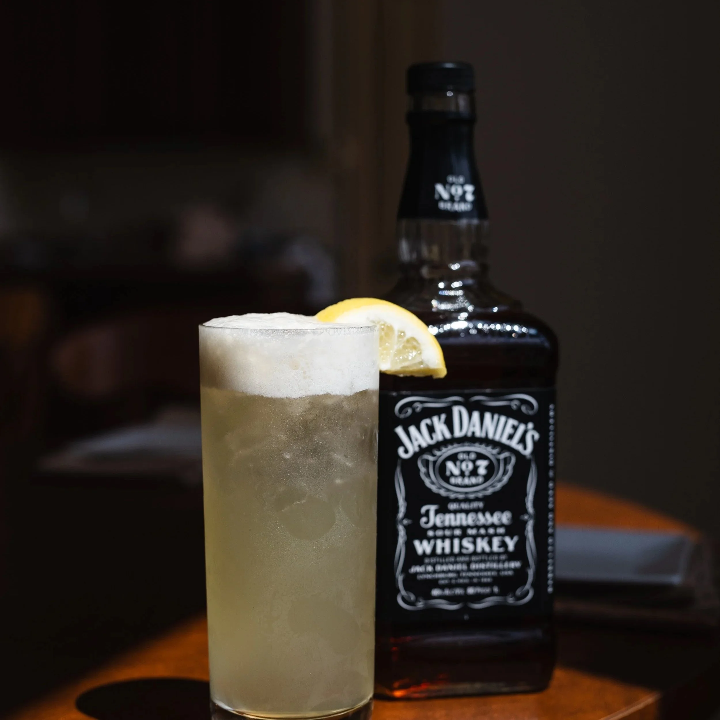 A glass of whiskey with a lemon wedge on the rim and a bottle of Jack Daniel's Tennessee whiskey in the background.