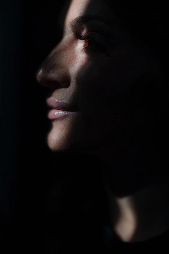 Close-up side profile of a woman's face, with soft lighting highlighting her lips, nose, and eye against a dark background.
