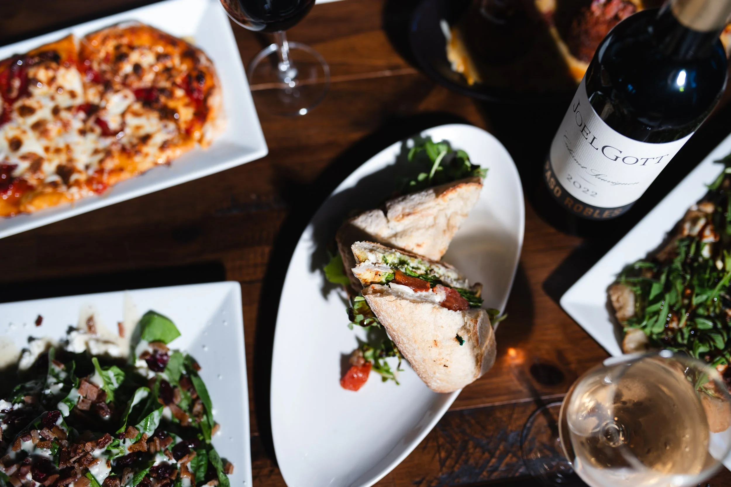 Assorted dishes including a pizza, sandwiches, salads, bottles of wine, and glasses of wine on a wooden table.