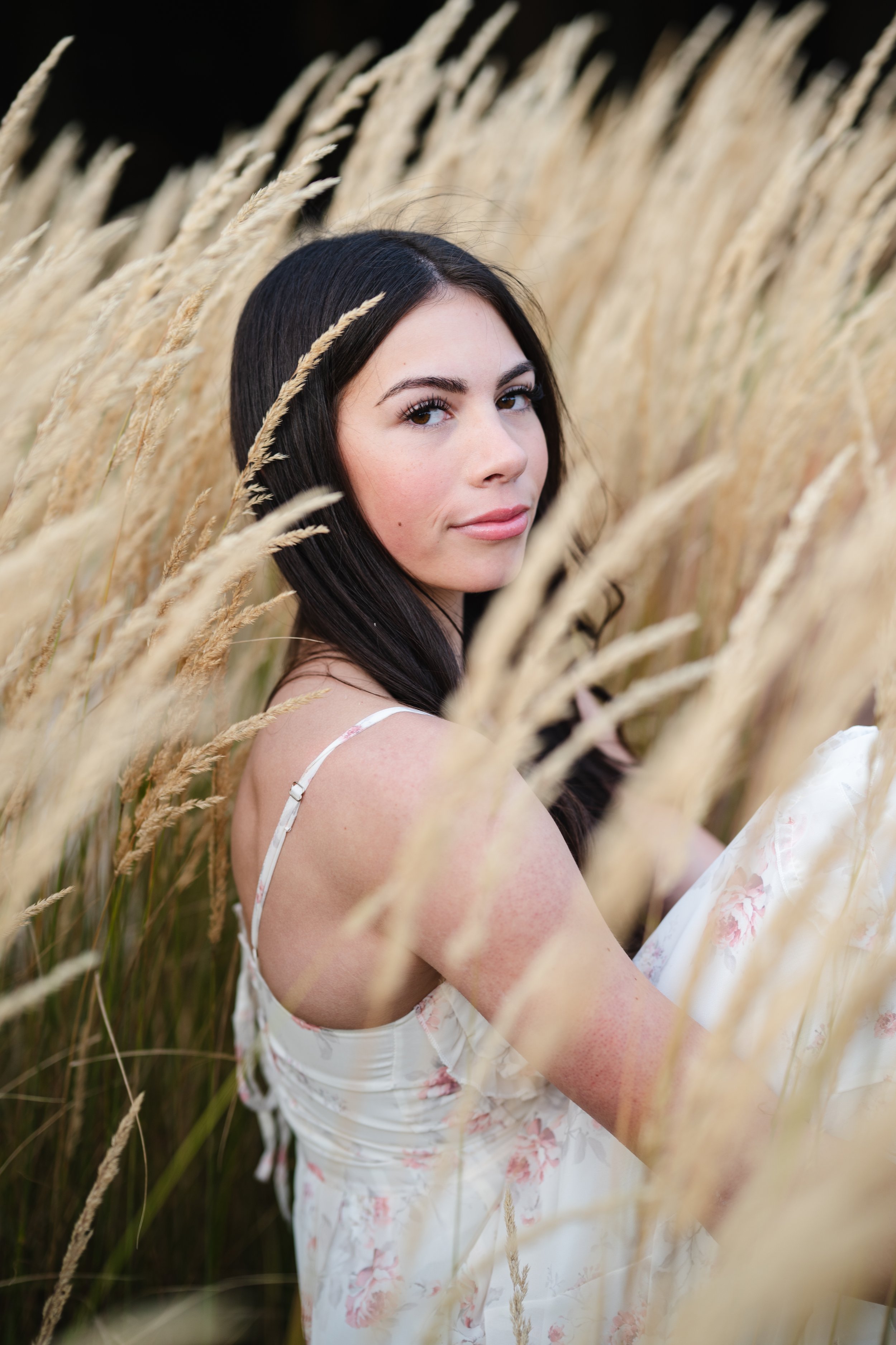 A young woman with long dark hair and light skin, wearing a floral dress, is standing in a field of tan grasses, looking over her shoulder with a slight smile.