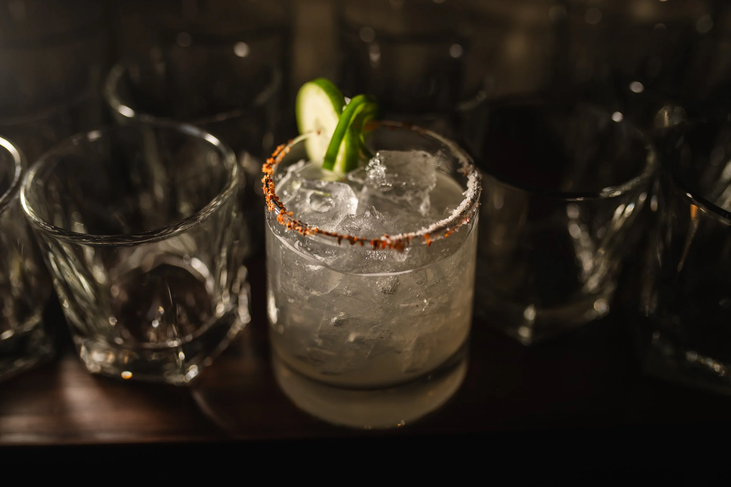 A close-up of a glass with a salted rim and lime wedge garnishing a cocktail, surrounded by empty shot glasses in a dimly lit setting.