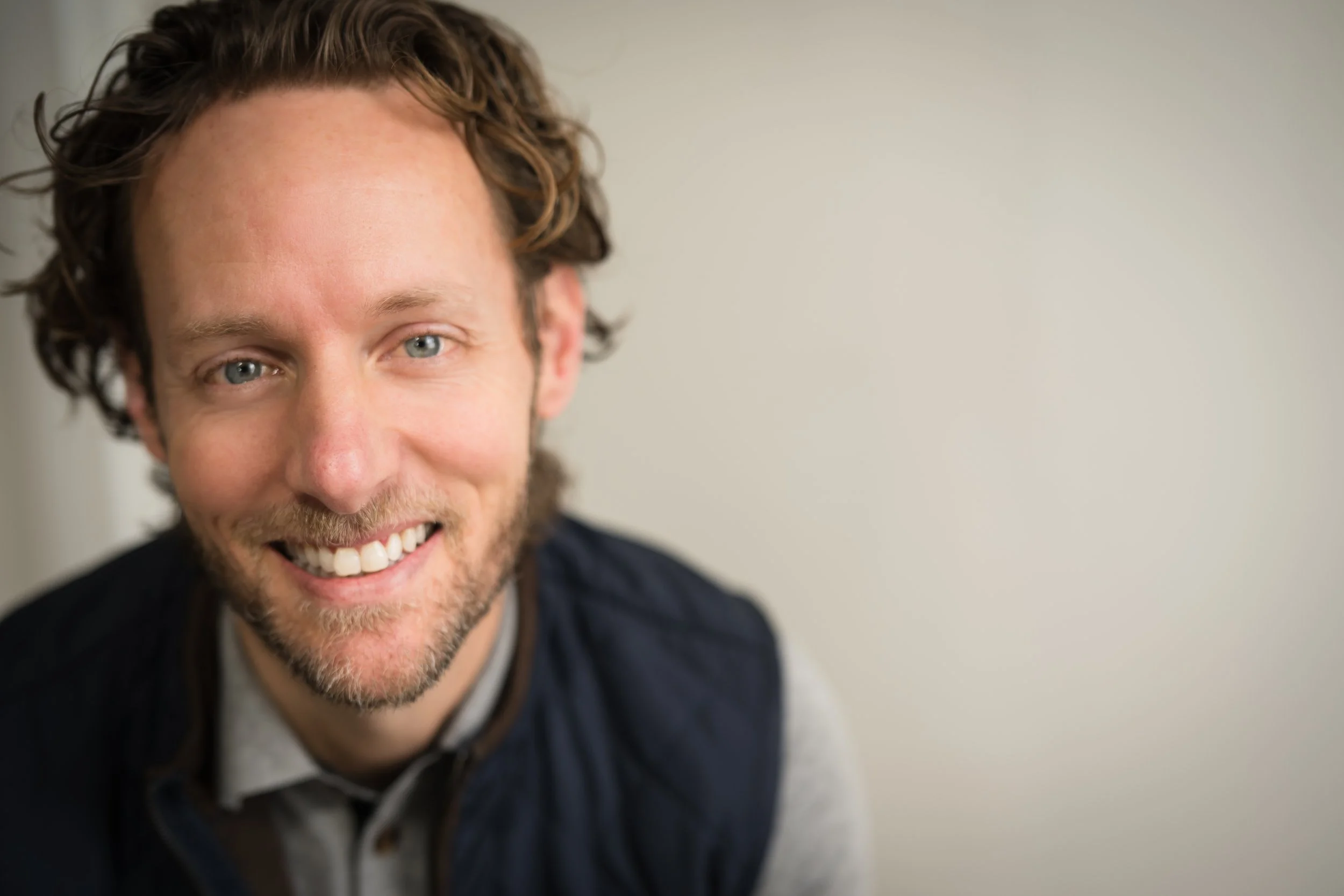 Paul Mattison, Photographer and owner A close-up of a smiling man with curly brown hair, blue eyes, and a beard, wearing a dark jacket and a gray shirt, against a plain background.