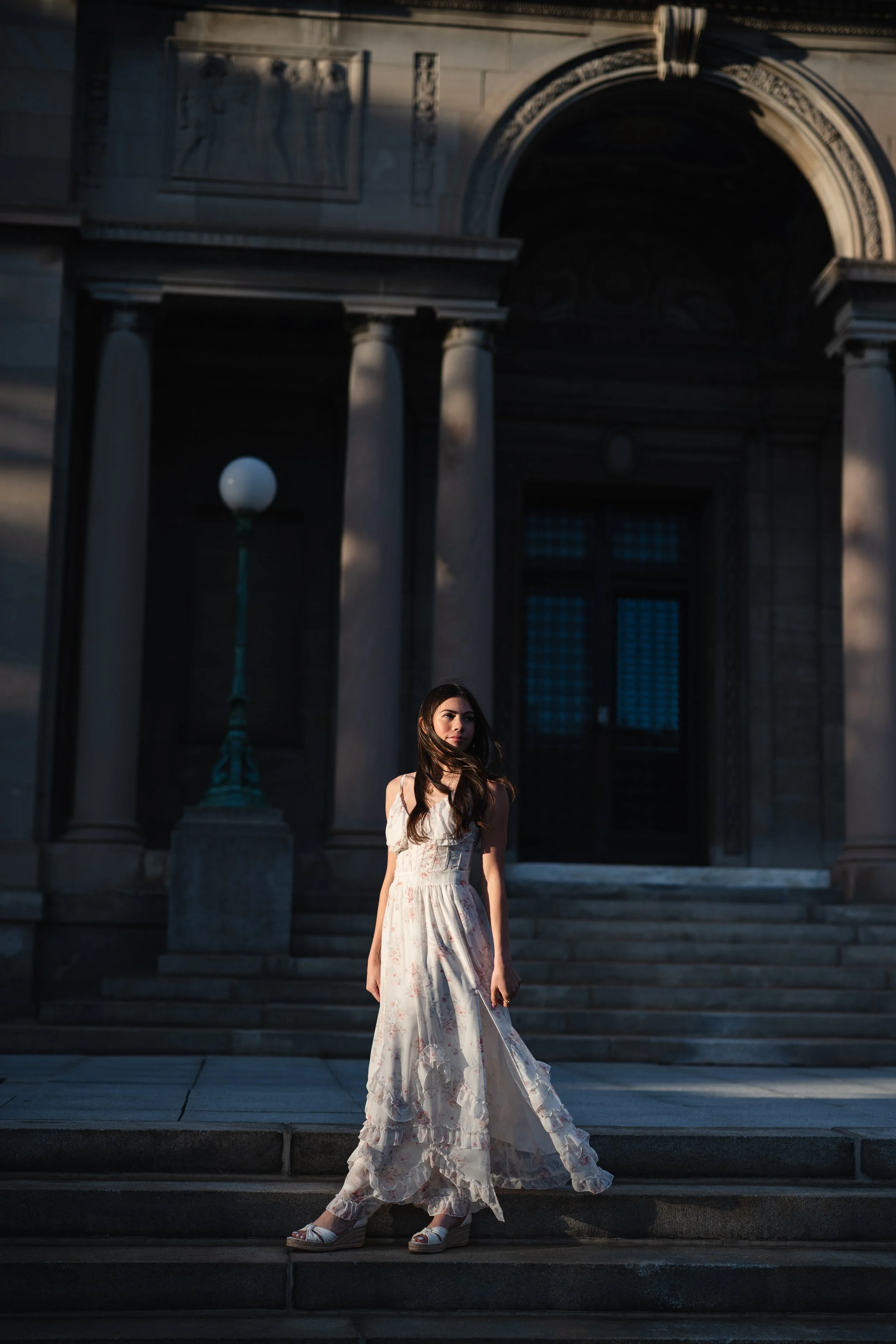 A young woman in a flowing white floral dress standing on a staircase in front of a historic building with columns at dusk.