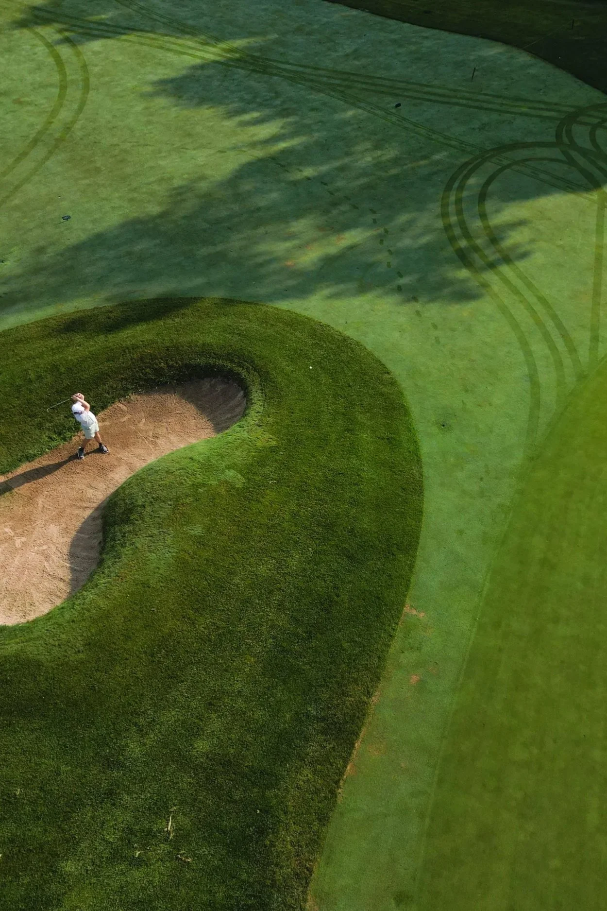 An aerial view of a golfer in white attire taking a shot from a sand bunker on a golf course, with well-maintained green grass and shadows cast by trees.