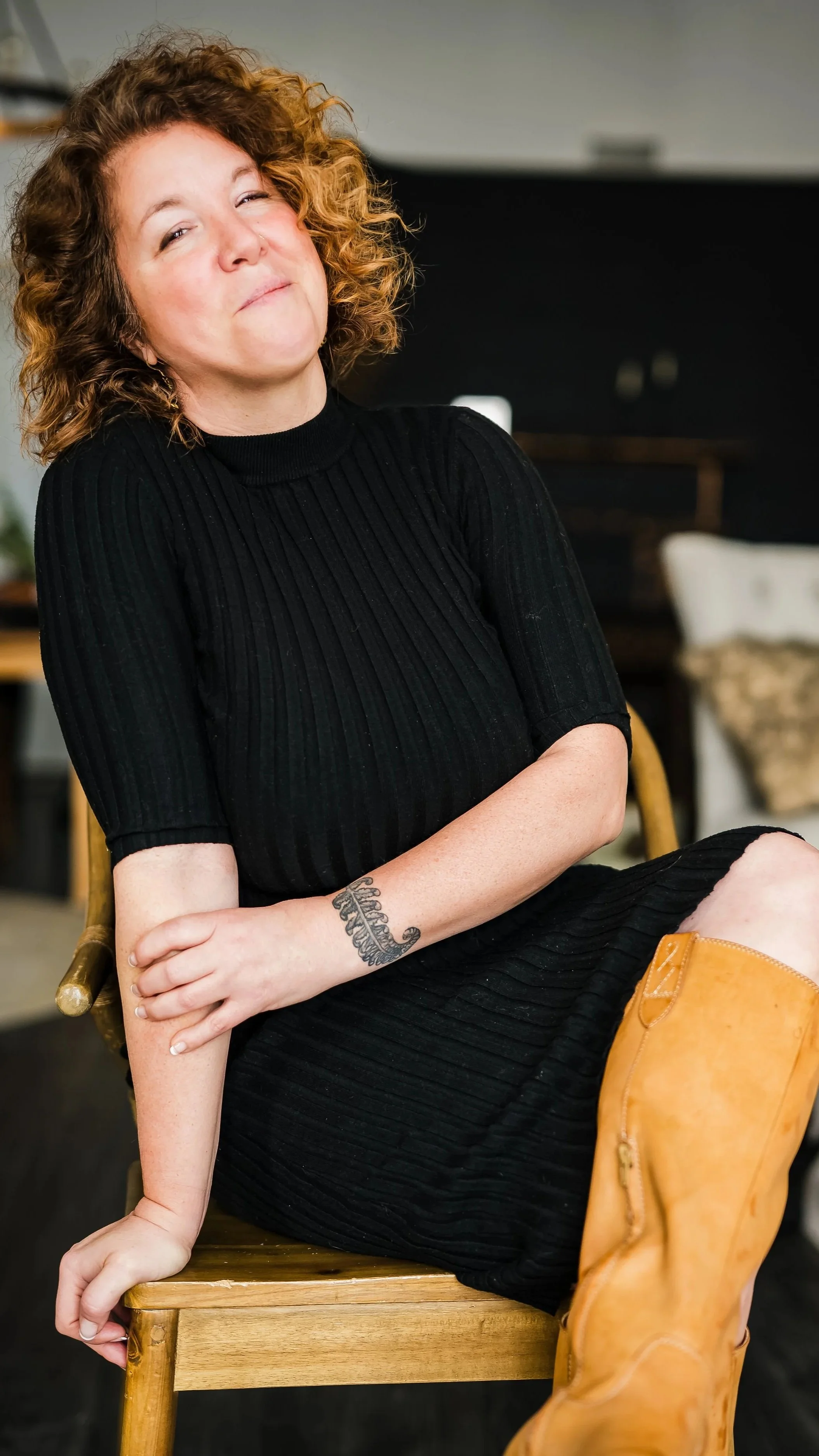Chelse Thompson, Photographer and owner, A woman with curly, reddish hair sitting on a wooden chair, wearing a black dress, with one arm resting on the chair and a tattoo on her arm, in an indoor setting.
