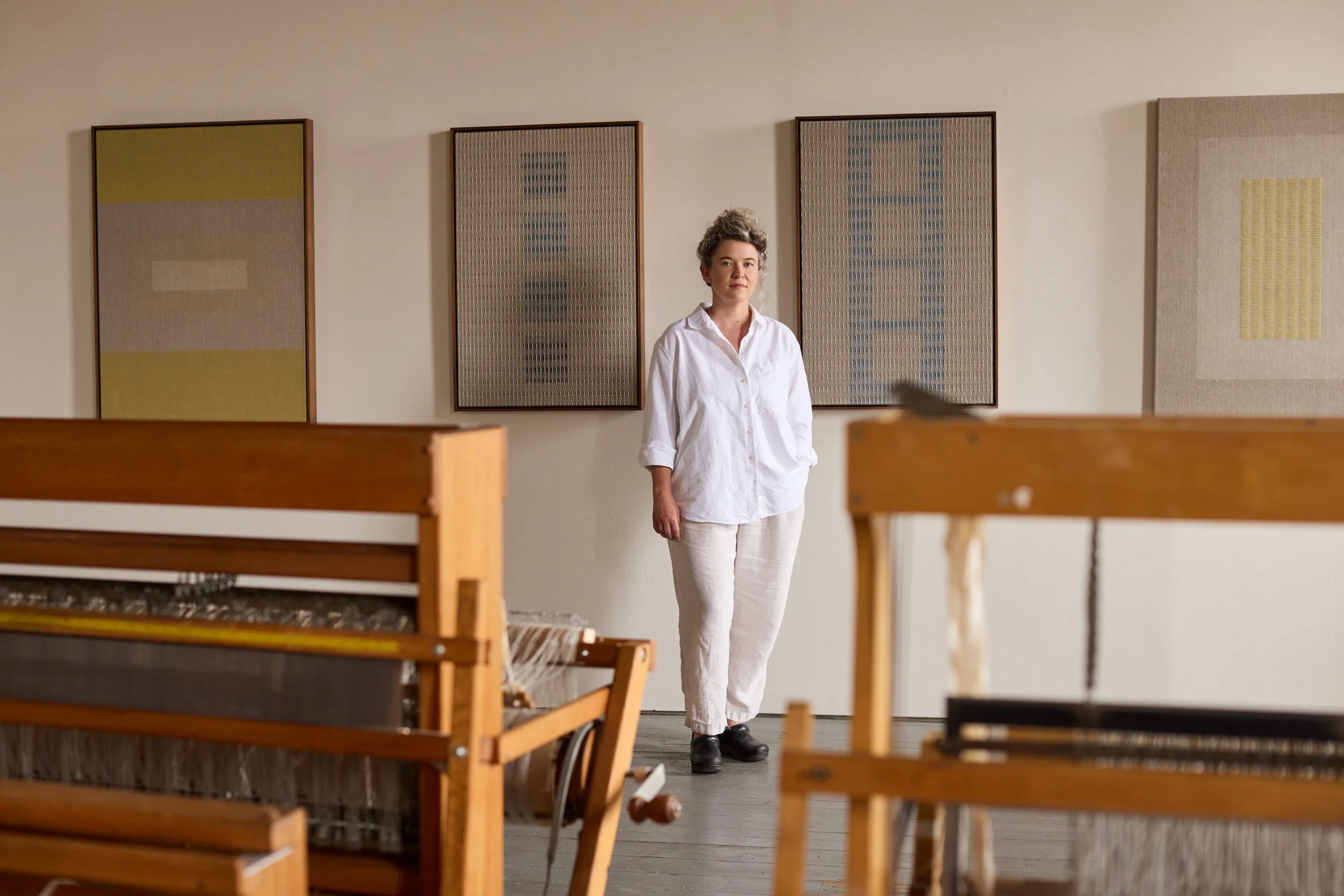 Portrait of Rachel DuVall standing in front of her woven artworks and behind her wood looms in her studio in Los Angeles.