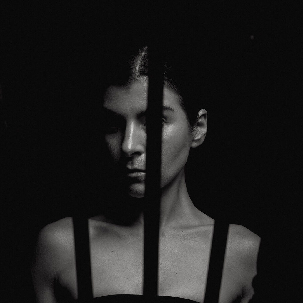 Black and white photo of a woman behind prison bars, with half her face obscured in shadow.