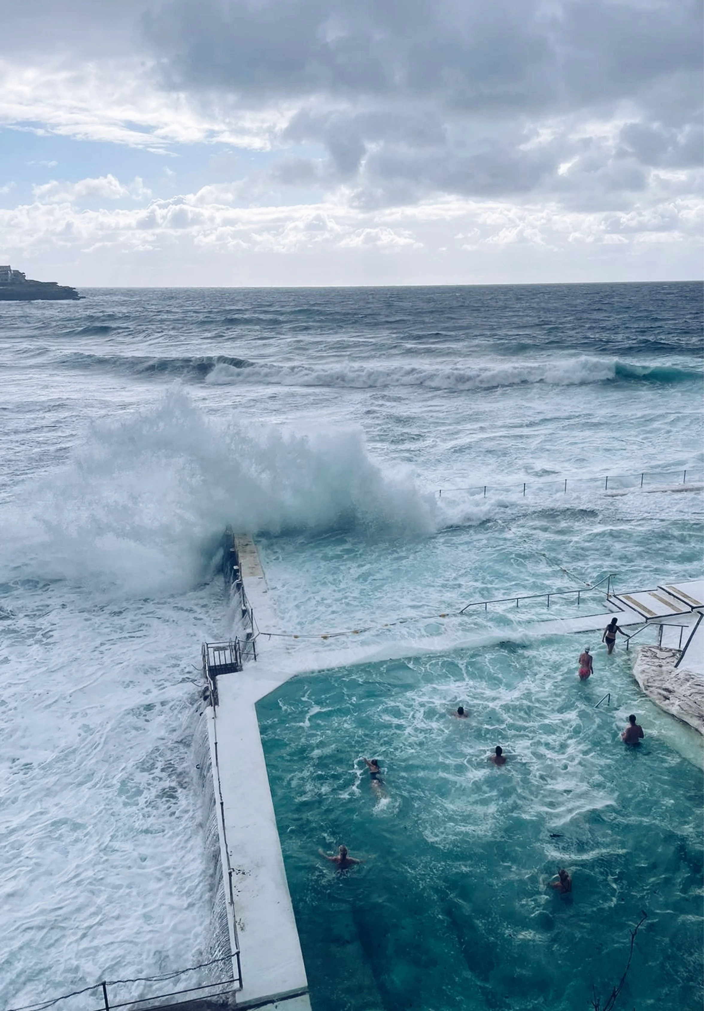People swimming in a pool next to the ocean, with waves crashing nearby and a cloudy sky overhead.